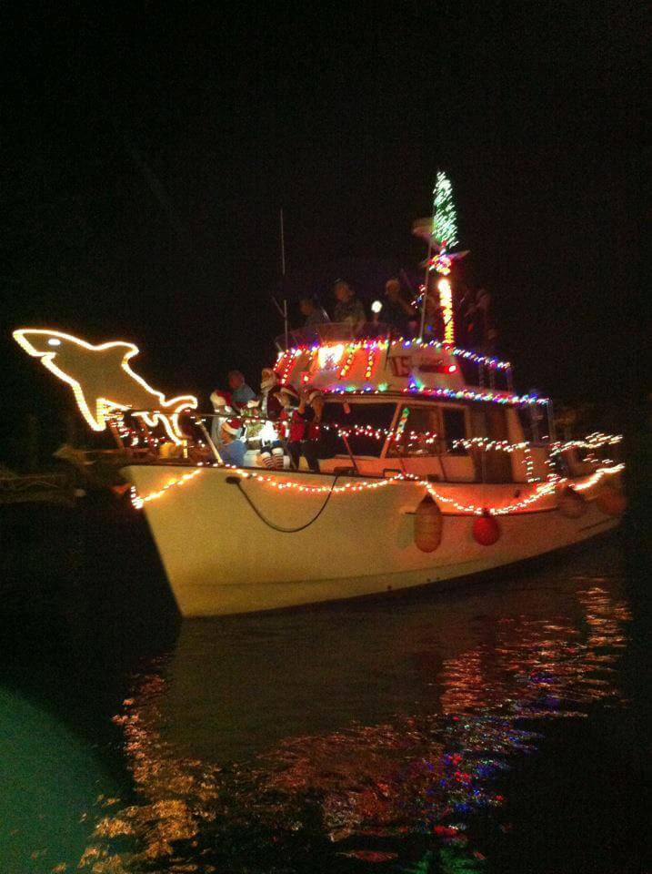 Boat decorated with Christmas lights. Partygoers can be seen in Christmas attire. There is a lit up shark on the bow.