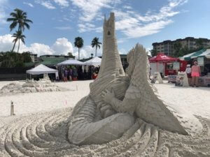 Sand sculpture of a large dragon in front of a tall castle tower.