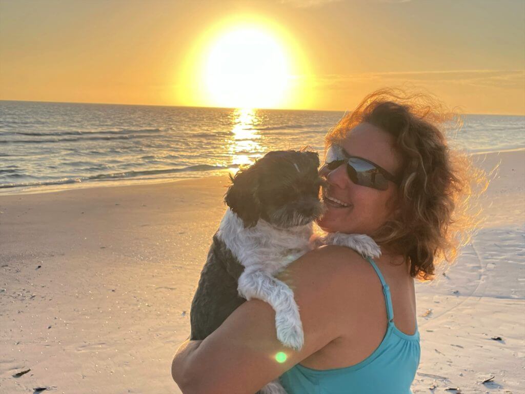 A woman holds her Shih Tzu on the beach as the sun sets.