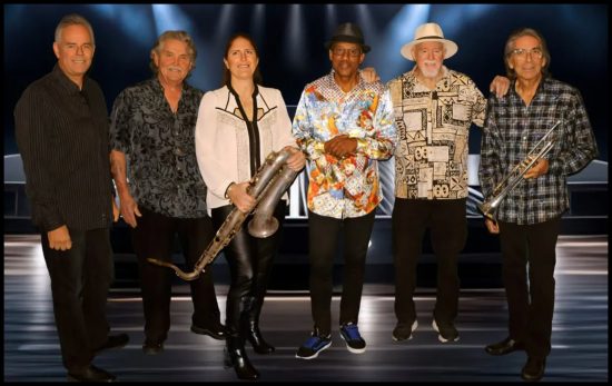 Six band members pose onstage with instruments, wearing colorful shirts under dramatic lighting.