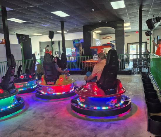 Indoor arcade ride with glowing LED spinning seats, patrons seated, and staff nearby under a high ceiling.
