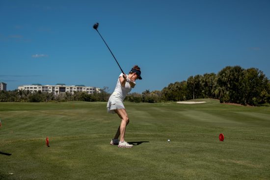 Golfer in mid‑swing on a sunny course with trees, sand bunker, and buildings in the background.