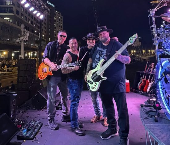Four band members pose on stage at night with guitars and drums, smiling toward the camera.