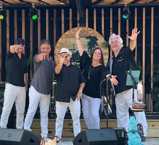 Five performers in black and white pose playfully on stage with tropical decor and music gear behind them.