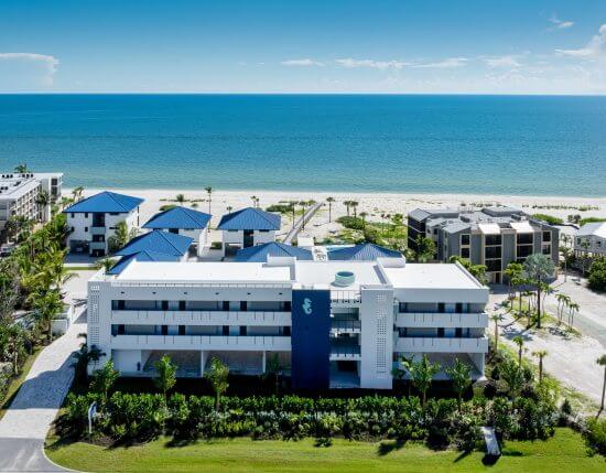Beachfront resort with white buildings, blue roofs, and a seahorse emblem facing a calm ocean and clear sky.