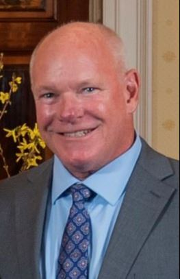 Man in grey suit, blue shirt, and patterned tie stands indoors near yellow flowers and wood paneling.