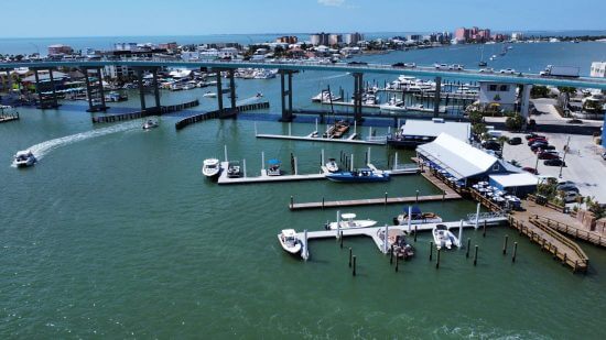 Marina with docked boats, a large bridge, and waterfront buildings along a calm coastal waterway.
