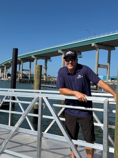 Person standing on a metal dock by the water with a large bridge in the background on a sunny day.