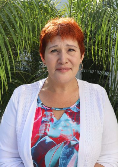 Person with short orange hair smiling outdoors, wearing a white cardigan and colorful top in front of green palm leaves.
