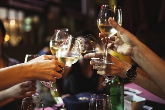 Hands clink glasses of white wine in a warm, lively restaurant setting during a social toast.