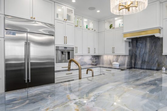 Luxurious white kitchen with marble counters, gold fixtures, glass cabinets, and a stainless steel fridge.