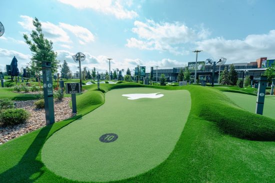 Mini‑golf course with green turf, mounds, palm trees, and a “Please Stay Off Mounds” sign under a blue sky.