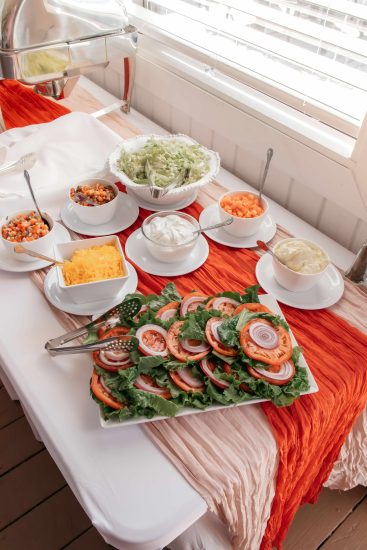 Salad bar setup with bowls of fresh toppings, including lettuce, tomatoes, cheese, onions, and sauces on a buffet table.