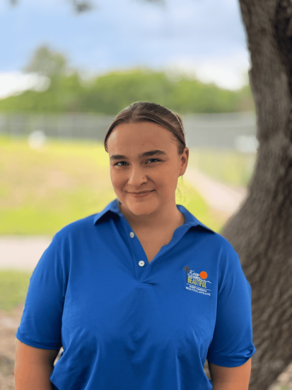 Person in bright blue “Keep Lee County Beautiful” shirt standing outdoors near a tree with greenery background.