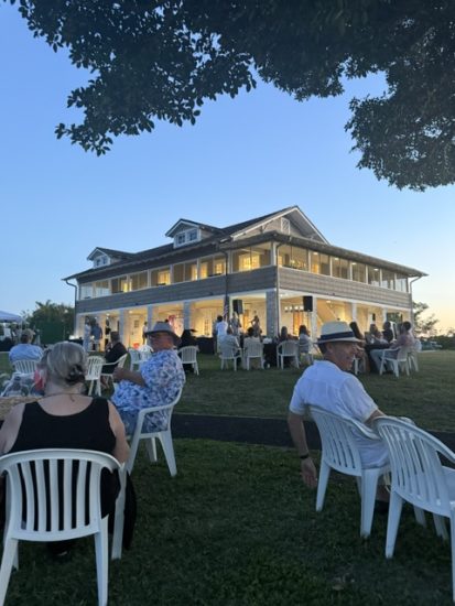 People seated and standing on a lawn at dusk in front of a large two‑story house with a wraparound porch and warm interior lighting.