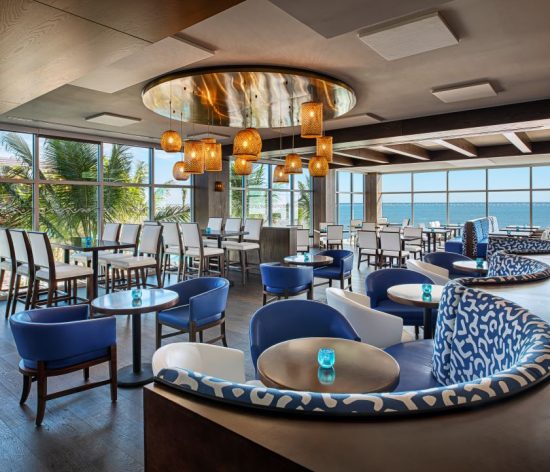 A bright, modern coastal‑style dining space with blue and white chairs, curved patterned booths, woven pendant lights, and large windows overlooking palm trees and the ocean.