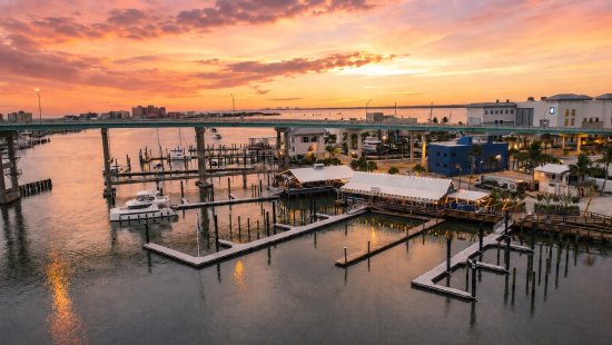 Aerial view of marina docks at sunset with bridge, buildings, and colorful orange sky reflecting on water.