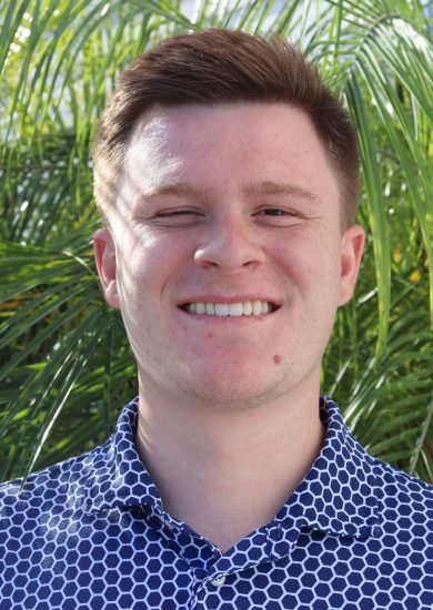 Person with short brown hair wearing a blue patterned shirt, standing outdoors in front of bright green palm leaves.