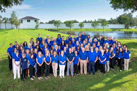 Large group of people in blue shirts posing outdoors near a pond with trees and a white building in the background.