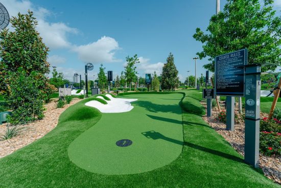 A landscaped miniature golf hole with bright green turf, a white sand-style bunker, rocks, shrubs, and signs marking course rules and Hole 1 of the black course.