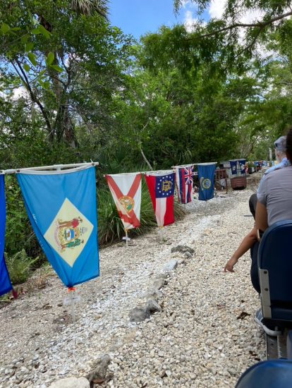 Passengers ride a small open-air train beside a gravel path lined with colorful state flags.
