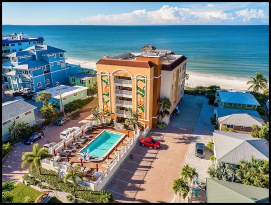 Aerial view of an oceanfront resort with orange murals, a pool, lounge chairs, and nearby pastel beach houses by the shoreline.