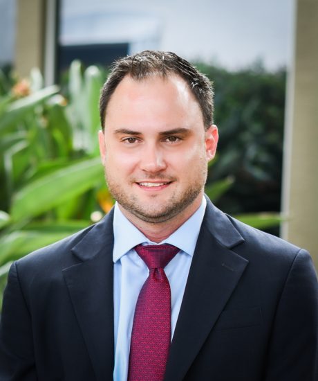 Man in a dark suit jacket, light blue dress shirt, and red patterned tie standing outdoors in front of green plants, smiling slightly.