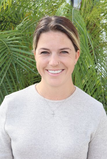 Portrait of a person standing outdoors in front of bright green palm leaves, wearing a light gray top and a small nose ring.