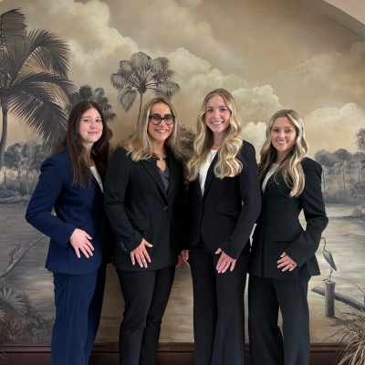 Four people in business suits standing in front of a painted mural of palm trees, water, and birds.