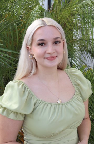 A person with long blonde hair in a light green puff‑sleeve dress standing outdoors in front of lush green plants.