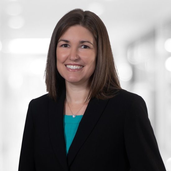 Woman with straight brown hair wearing a black blazer and teal top, posing in a softly blurred indoor professional setting.