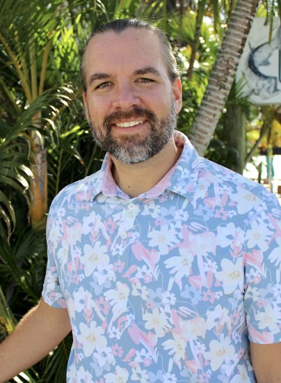 Person with long hair and a beard stands outdoors in front of lush tropical plants, wearing a light blue floral shirt.