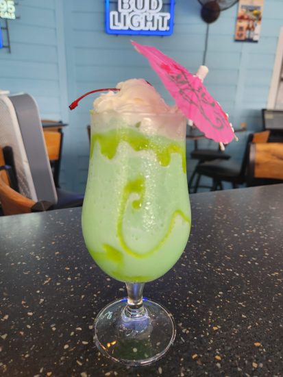 Tropical green cocktail with whipped cream, cherry, and umbrella on a bar counter beneath a Bud Light sign.