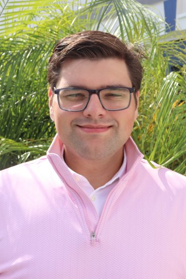 Man in glasses wearing a light pink quarter‑zip over a white collared shirt, standing outdoors by palm leaves in bright sunlight.