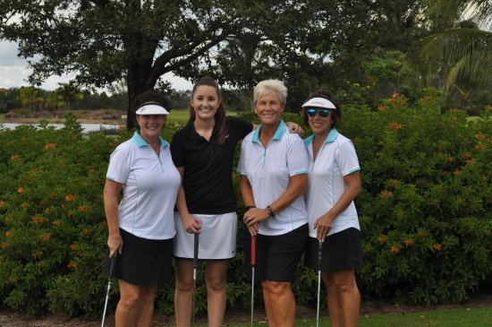 Four women in golf attire pose smiling on a golf course, holding clubs, with lush greenery and orange flowers behind them.