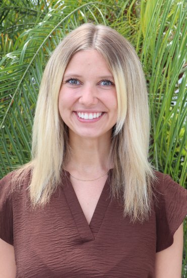 Woman with shoulder‑length blonde hair standing outdoors in front of bright green palm leaves, wearing a brown textured top and a thin gold necklace.