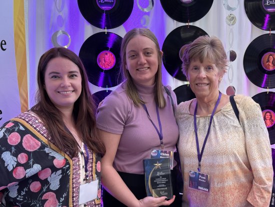 Three women smile with an award trophy at an event with vinyl record backdrop.
