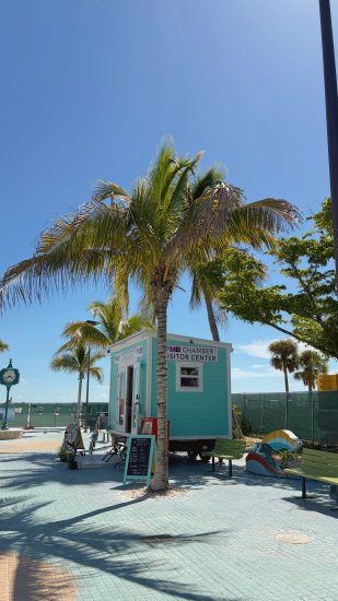 FMB Chamber Visitor Center turquoise kiosk under palm trees on sunny boardwalk with green benches and clear blue sky.